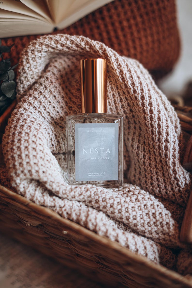 May include: A rectangular glass perfume bottle with a gold cap, labeled "NESTA LEATHER & STEEL", rests on a beige knitted blanket inside a woven basket. The bottle contains 50 ml of perfume. The background includes a book and more knitted items.