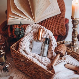 May include: A woven basket holds two perfume bottles, one square and one cylindrical, both labeled "NESTA". The basket rests on a cream-colored knitted blanket, with a stack of books and a lit candle in the background. A wooden mushroom-shaped object is also in the basket.