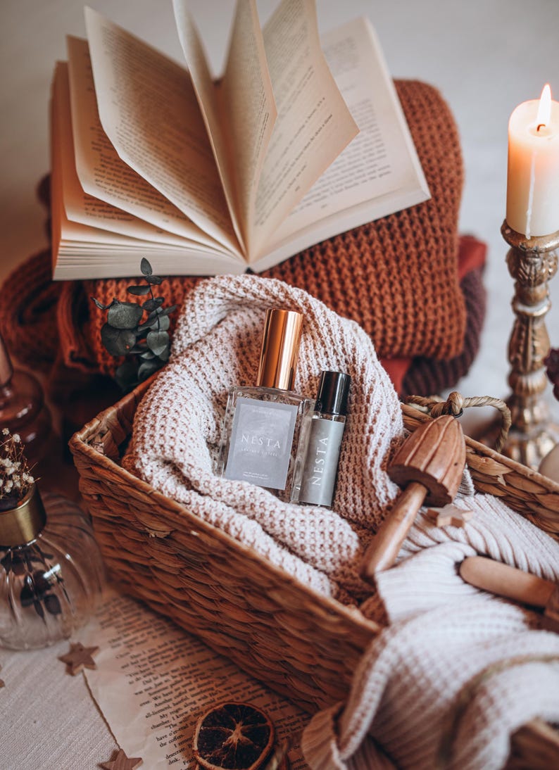 May include: A woven basket holds two perfume bottles, one square and one cylindrical, both labeled "NESTA". The basket rests on a cream-colored knitted blanket, with a stack of books and a lit candle in the background. A wooden mushroom-shaped object is also in the basket.