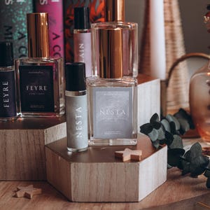 May include: A collection of perfume bottles with gold-colored caps and labels, arranged on wooden blocks. The bottles are labeled "Feyre" and "Nesta". Books by Sarah J. Maas are in the background, along with a lit candle.