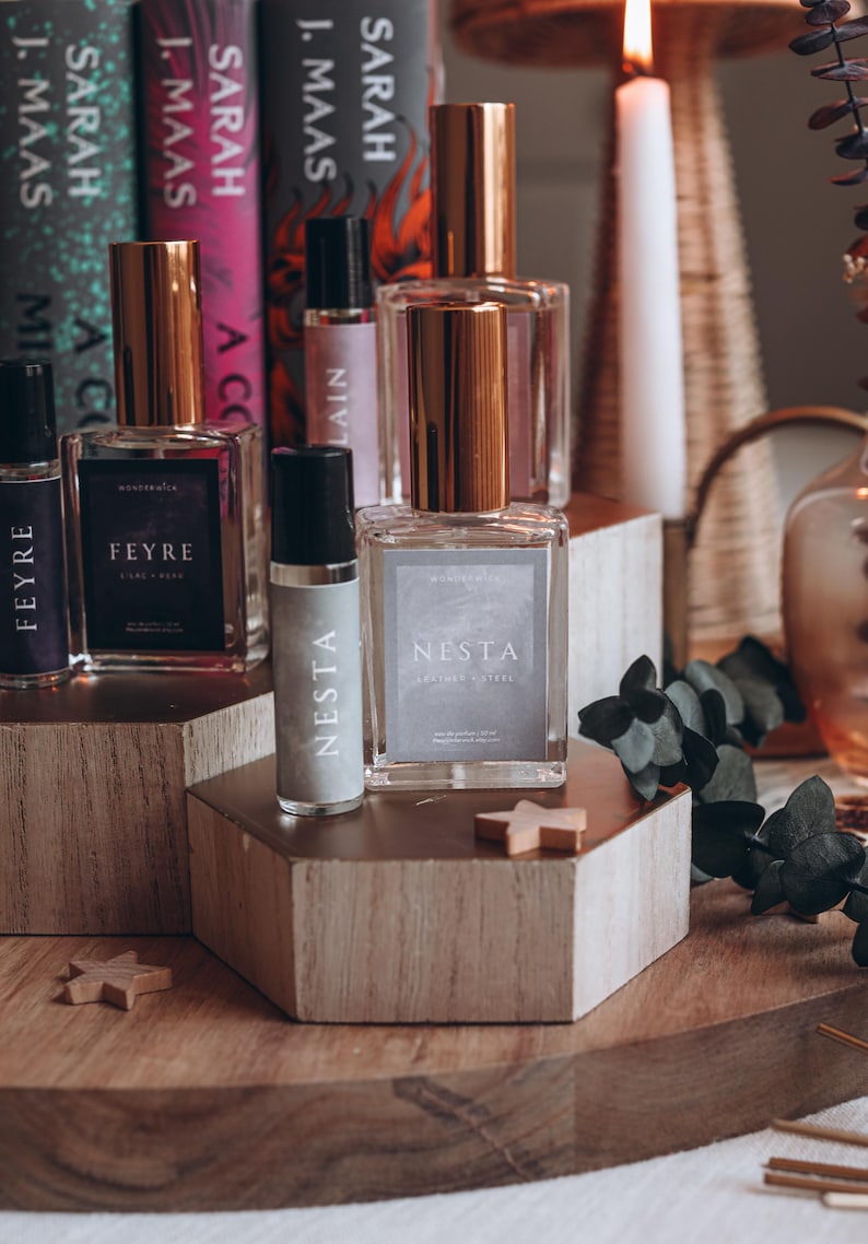 May include: A collection of perfume bottles with gold-colored caps and labels, arranged on wooden blocks. The bottles are labeled "Feyre" and "Nesta". Books by Sarah J. Maas are in the background, along with a lit candle.