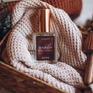 May include: A perfume bottle with a gold cap and a dark burgundy label that reads "MANON, Plum + Light Musk" sits in a woven basket on a cream-coloured knitted blanket. The bottle is from Wonderwick, with a 50 ml size.