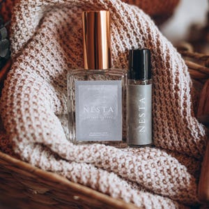 May include: Two perfume bottles, one large and one small, sit in a woven basket atop a knitted blanket. The larger bottle has a gold cap and a label that reads "NESTA LEATHER + STEEL". The smaller bottle has a black cap and a label that reads "NESTA".