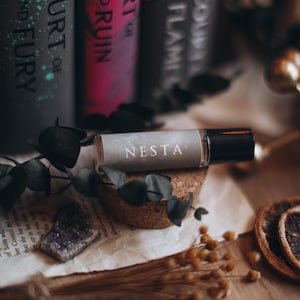 May include: A roll-on essential oil bottle labeled "NESTA" rests on a cork base, with a backdrop of books and dried botanicals. The bottle is clear with a black cap. The books have titles like "A Court of Mist and Fury".