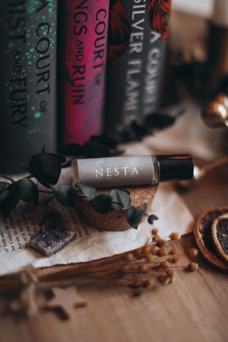 May include: A roll-on essential oil bottle labeled "NESTA" rests on a cork base, with a backdrop of books and dried botanicals. The bottle is clear with a black cap. The books have titles like "A Court of Mist and Fury".