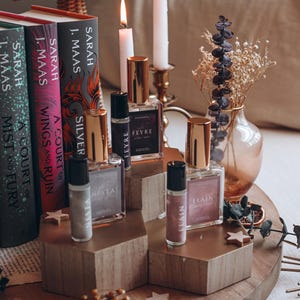 May include: A display of perfume bottles and rollerballs with gold caps, arranged on wooden platforms. The bottles are labeled "FEYRE", "ELAIN", and "NESTA". Books by Sarah J. Maas are in the background, along with candles and decorative elements.