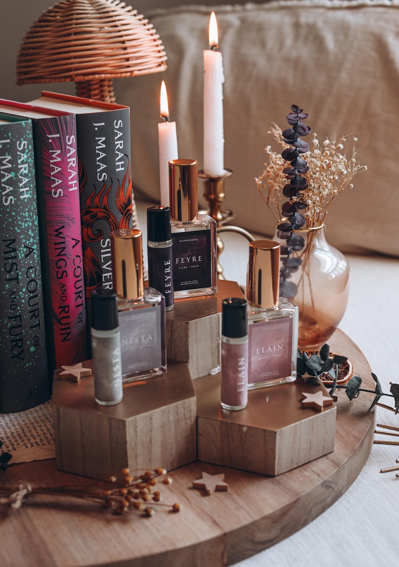 May include: A display of perfume bottles and rollerballs with gold caps, arranged on wooden platforms. The bottles are labeled "FEYRE", "ELAIN", and "NESTA". Books by Sarah J. Maas are in the background, along with candles and decorative elements.