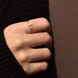 Könnte beinhalten: Ein zarter Goldring mit einem kleinen, mit Diamanten besetzten Kreuzdesign.