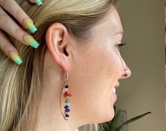 May include: Copper-colored wire earrings with a vertical row of colorful gemstones. The gemstones include red, orange, yellow, green, blue, indigo, and violet. The earrings are worn by a person with blonde hair.