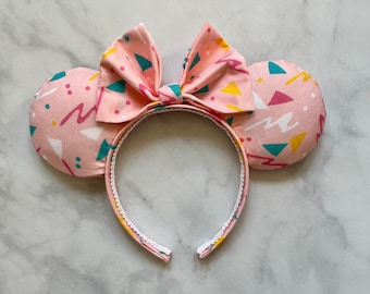 May include: Pink headband with large, fabric-covered ears featuring a geometric pattern in pink, yellow, and teal. The headband has a white bow in the center.