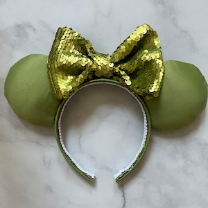 Puede incluir: Orejas de ratón de satén verde con un lazo de lentejuelas doradas. La diadema es blanca con un ribete verde.