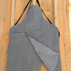 May include: A black and white striped apron with a large pocket on the front. The apron is hanging on a wooden wall.