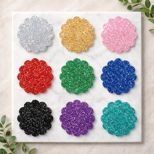 May include: A set of nine circular coasters with scalloped edges, each featuring a distinct glittery finish in silver, gold, pink, red, green, blue, black, purple, and teal, arranged in a three-by-three grid on a white marble surface.