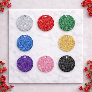 May include: Nine glittery, circular tags in various colors: silver, red, green, blue, gold, purple, pink, and black. Each tag has a small hole at the top for hanging. The tags are arranged on a white marble surface.