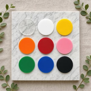 May include: A set of nine circular acrylic discs arranged in a three-by-three grid. The collection includes one clear disc, one white, one yellow, one orange, one red, one pink, one green, one blue, and one black disc, all with a smooth finish.
