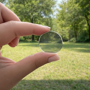 May include: A clear, circular lens is held between fingers against a backdrop of green grass and trees. The lens is transparent, reflecting the natural light and surroundings. The image is taken outdoors on a sunny day.