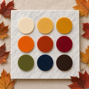 May include: Nine circular discs arranged in a three-by-three grid on a square white marble slab. The discs feature a smooth, matte finish in various colors including cream, yellow, mustard, orange, rust, burgundy, olive green, navy, and brown.