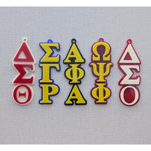 May include: Five colorful Greek letter pendants in red, yellow, blue, and purple. Each pendant features a different combination of Greek letters, with a small hole at the top for hanging. The pendants are arranged in a row against a light gray background.