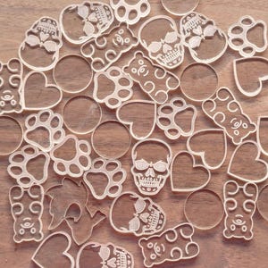 May include: A collection of clear acrylic charms in various shapes including skulls, teddy bears, paw prints, hearts, and circles. These transparent, flat decorative pieces feature detailed outlines and are arranged in a scattered pattern.