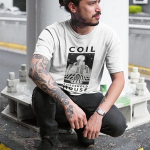 May include: A white t-shirt featuring a black and white graphic of a building with the text "COIL THRESHOLD HOUSE".
