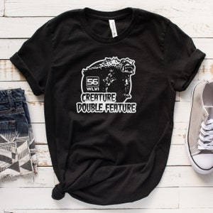 以下が含まれることがあります： モンスターと「Creature Double Feature」と「56 WLVI」の文字が白で描かれた黒いTシャツ。