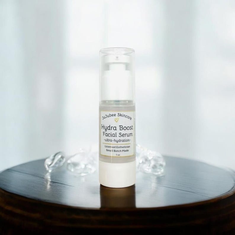 Pu&ograve; includere: Flacone bianco e trasparente di siero viso Hydra Boost di JuJubee Skincare con dosatore a pompa. L'etichetta include il testo "ultra-hydration" e "1 oz". Il flacone &egrave; su una superficie di legno scuro.