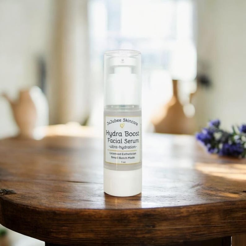 Pu&ograve; includere: Una bottiglia trasparente e bianca di siero viso Hydra Boost di JuJube Skincare &egrave; appoggiata su un tavolo di legno. La bottiglia ha un dosatore a pompa e un'etichetta con informazioni sul prodotto. Lo sfondo &egrave; sfocato.