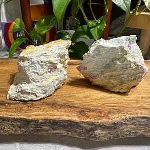 May include: Two rough, light grey and white rocks with pink crystals embedded in them. The rocks are sitting on a wooden surface.