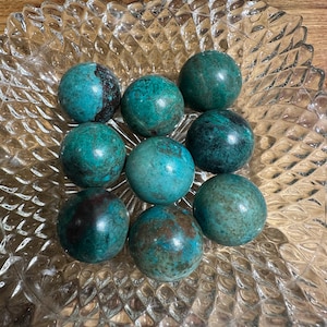 May include: Eight polished turquoise spheres with dark brown and green markings are displayed in a clear glass dish. The spheres are arranged in a clear, decorative glass dish with a diamond pattern. The dish sits on a wooden surface.
