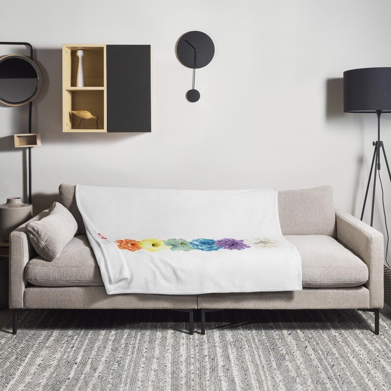 May include: A white throw blanket with a rainbow floral design. The blanket is draped over a light gray couch with a pillow. The couch is in a living room setting with a floor lamp and a clock on the wall.