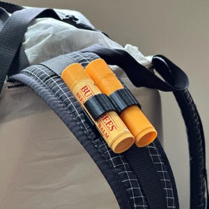 May include: Close-up of a backpack strap with two orange lip balm tubes secured by a black holder. One tube displays the text "Burt's Bees Balm" in a red and yellow label. The backpack has a black and white checkered pattern.