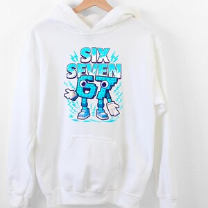 May include: White hooded sweatshirt with a front graphic. The graphic displays the words "SIX SEVEN" in blue, with the numbers "67" designed as cartoon characters with arms, legs, and shoes. The design has a playful, cartoonish style.