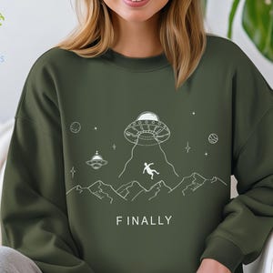 K&ouml;nnte beinhalten: Gr&uuml;nes Sweatshirt mit einer wei&szlig;en Grafik eines UFOs, einer Person, die aus dem UFO f&auml;llt, und Bergen. Der Text "FINALLY" ist unter der Grafik gedruckt.