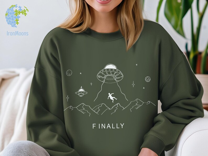 K&ouml;nnte beinhalten: Gr&uuml;nes Sweatshirt mit einer wei&szlig;en Grafik eines UFOs, einer Person, die aus dem UFO f&auml;llt, und Bergen. Der Text "FINALLY" ist unter der Grafik gedruckt.