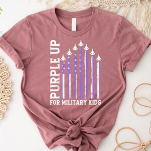 May include: A mauve t-shirt with a purple up for military kids graphic. The graphic features a purple and white American flag with airplanes flying over it.