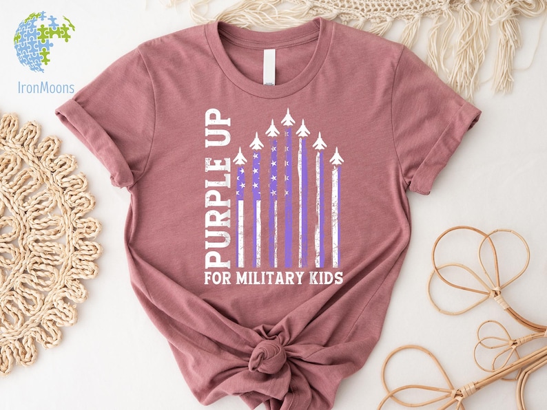 May include: A mauve t-shirt with a purple up for military kids graphic. The graphic features a purple and white American flag with airplanes flying over it.