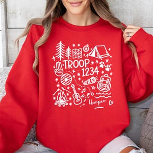 May include: A red sweatshirt with a white graphic design featuring camping-themed illustrations, the text "TROOP 1234", and the name "Harper". The design includes trees, a tent, a compass, a campfire, a guitar, and a backpack.