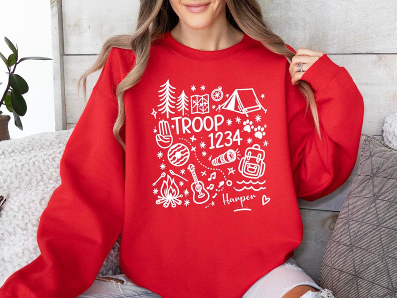 May include: A red sweatshirt with a white graphic design featuring camping-themed illustrations, the text "TROOP 1234", and the name "Harper". The design includes trees, a tent, a compass, a campfire, a guitar, and a backpack.