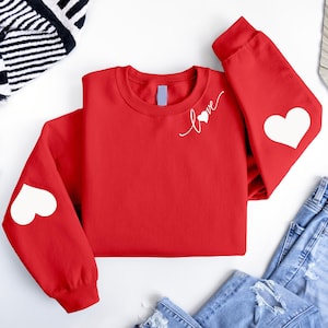 May include: Red sweatshirt with white hearts on the elbows and the word "love" with a heart on the chest.