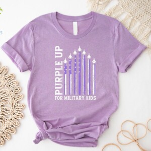 May include: Purple Up for Military Kids t-shirt in a lavender color with a white American flag design featuring purple stripes and airplanes.
