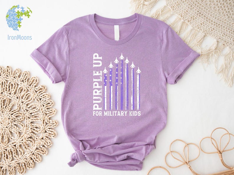 May include: Purple Up for Military Kids t-shirt in a lavender color with a white American flag design featuring purple stripes and airplanes.