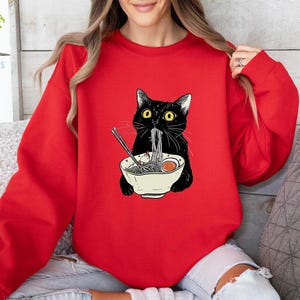 Peut inclure: Un sweat-shirt rouge avec un chat noir mangeant des nouilles ramen. Le chat porte un collier noir et a les yeux verts. Le bol de ramen est blanc avec un bord rouge.