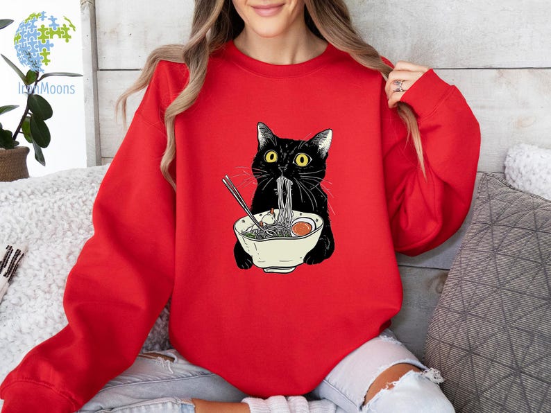 Peut inclure: Un sweat-shirt rouge avec un chat noir mangeant des nouilles ramen. Le chat porte un collier noir et a les yeux verts. Le bol de ramen est blanc avec un bord rouge.