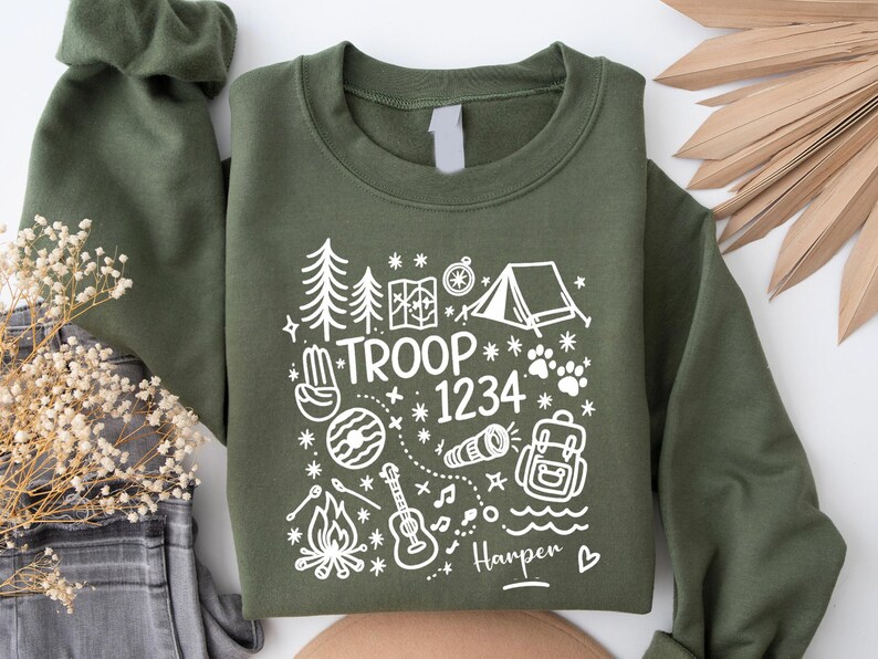 May include: Olive green sweatshirt with white graphic of camping elements, including trees, a tent, and a map. The text "TROOP 1234" and the name "Harper" are also featured. A cozy and stylish top for outdoor enthusiasts.