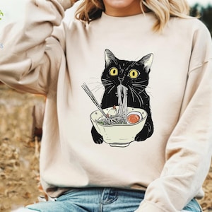 Peut inclure: Un sweat-shirt blanc avec un chat noir mangeant des nouilles ramen. Le chat a les yeux jaunes et porte un bol sur la t&ecirc;te. Les nouilles ramen sont dans un bol blanc avec des baguettes.