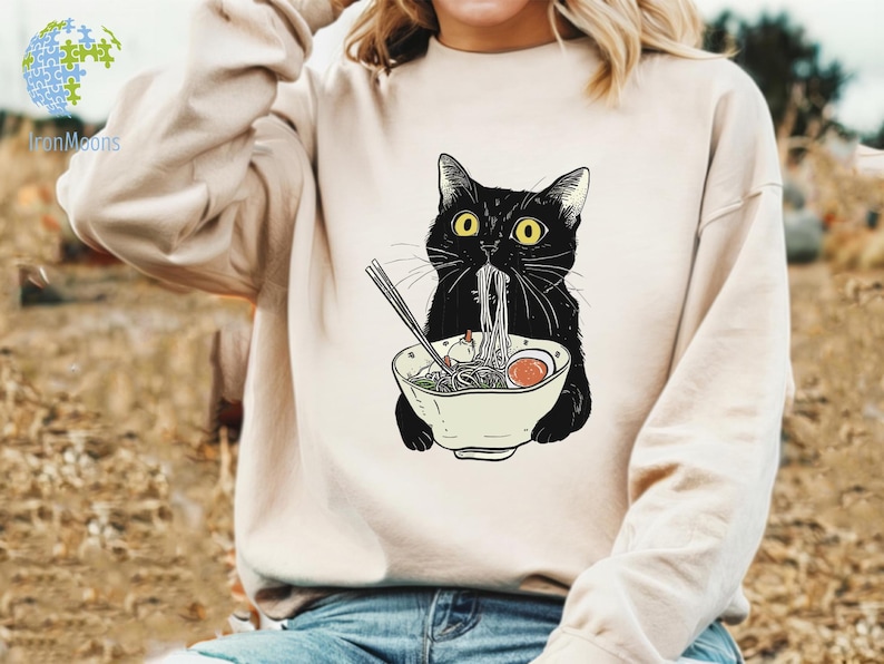 Peut inclure: Un sweat-shirt blanc avec un chat noir mangeant des nouilles ramen. Le chat a les yeux jaunes et porte un bol sur la t&ecirc;te. Les nouilles ramen sont dans un bol blanc avec des baguettes.