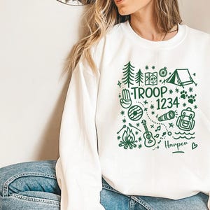 May include: White sweatshirt with a green graphic design featuring camping-themed illustrations and the text "TROOP 1234" along with a name. The design includes trees, a tent, a compass, and a guitar.
