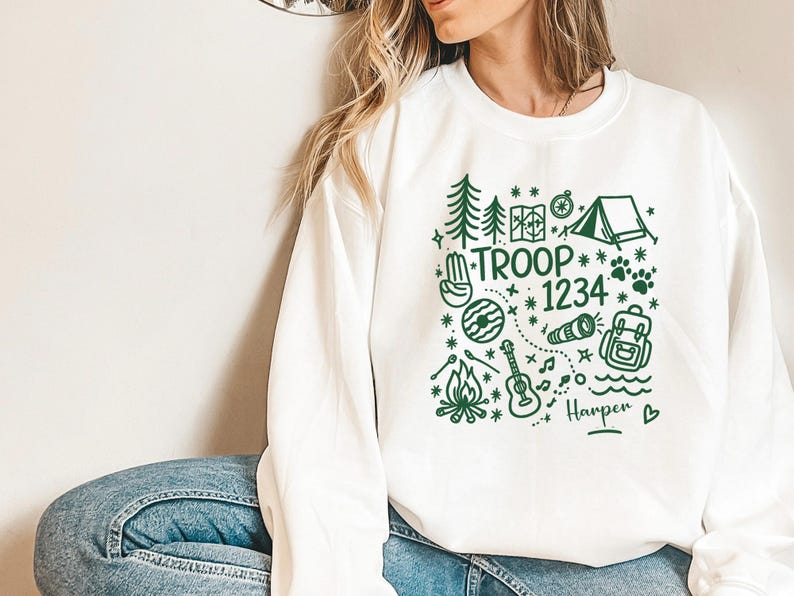 May include: White sweatshirt with a green graphic design featuring camping-themed illustrations and the text "TROOP 1234" along with a name. The design includes trees, a tent, a compass, and a guitar.