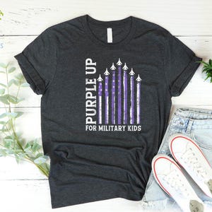 May include: A dark gray t-shirt with a purple up for military kids design. The design features a purple American flag with white airplanes flying over it.