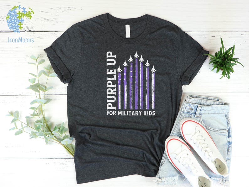 May include: A dark gray t-shirt with a purple up for military kids design. The design features a purple American flag with white airplanes flying over it.